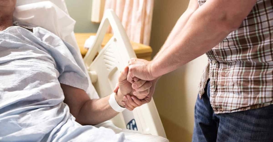 A man holding the hand of an older person who is resting in a hospital bed