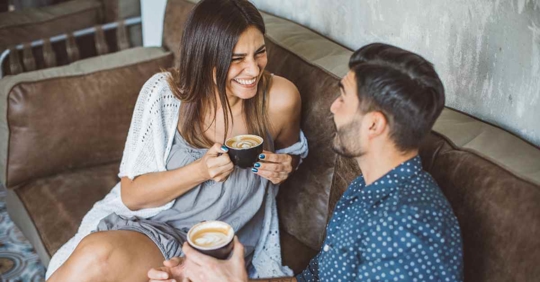 A man and woman sitting on a couch drinking coffee. They are both smiling and laughing