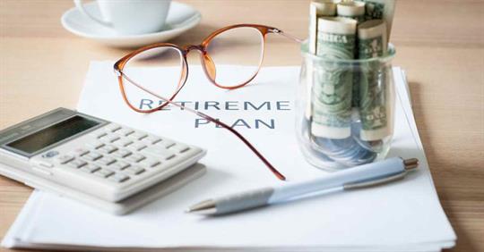 Glasses, a calculator, a pen, and money resting on a pile of paper that reads "retirement plan"